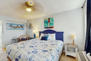 A hotel room with a bed, a nightstand, a chair, a painting of palm trees, and a painting of a lighthouse. - Palms Resort Unit 404 (Myrtle Beach)
