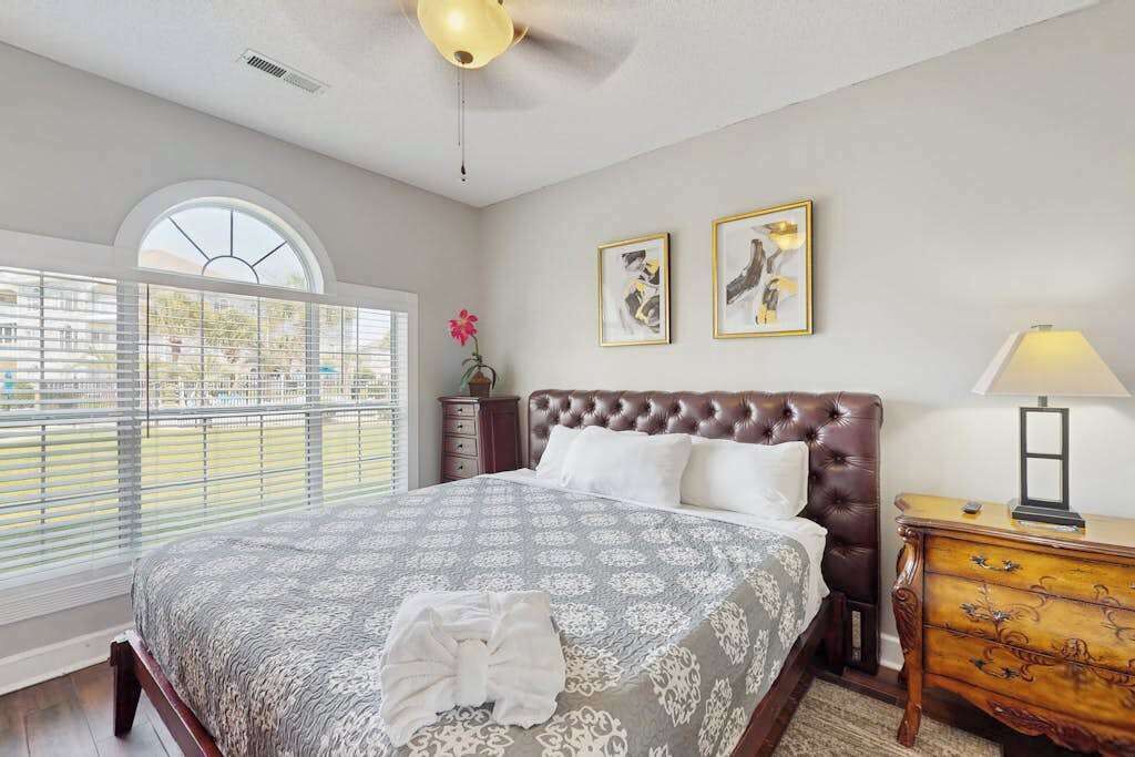 A bedroom with a large bed, a window with blinds, a ceiling fan, and two framed pictures on the wall.