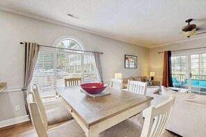 A dining area with a wooden table, white chairs, and a red bowl. There is a window with blinds, a sofa, and a ceiling fan. - The Iris | One Oh One (Myrtle Beach)