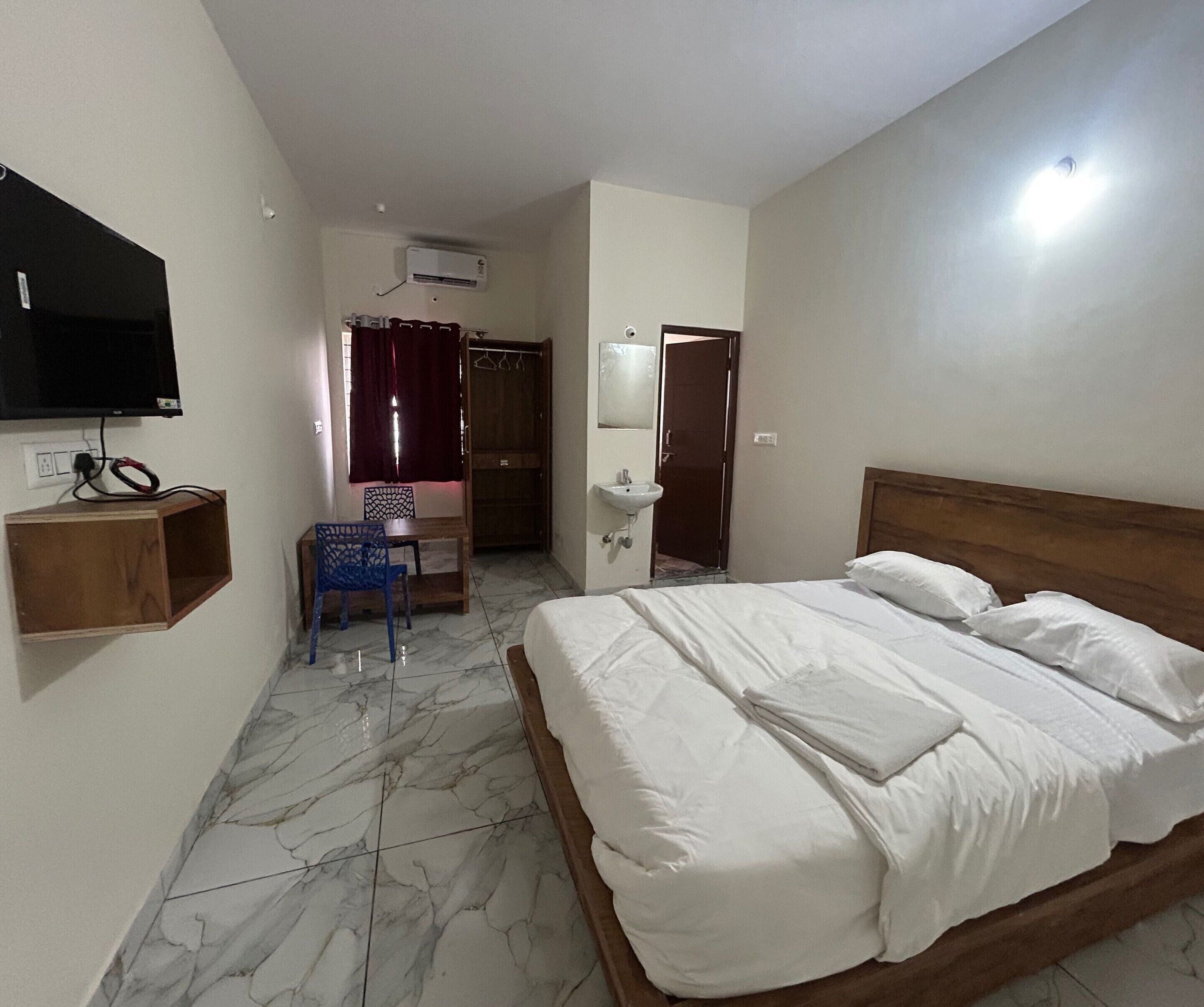A hotel room with a bed, a TV mounted on the wall, a small table with a chair, and a bathroom with a mirror and sink.