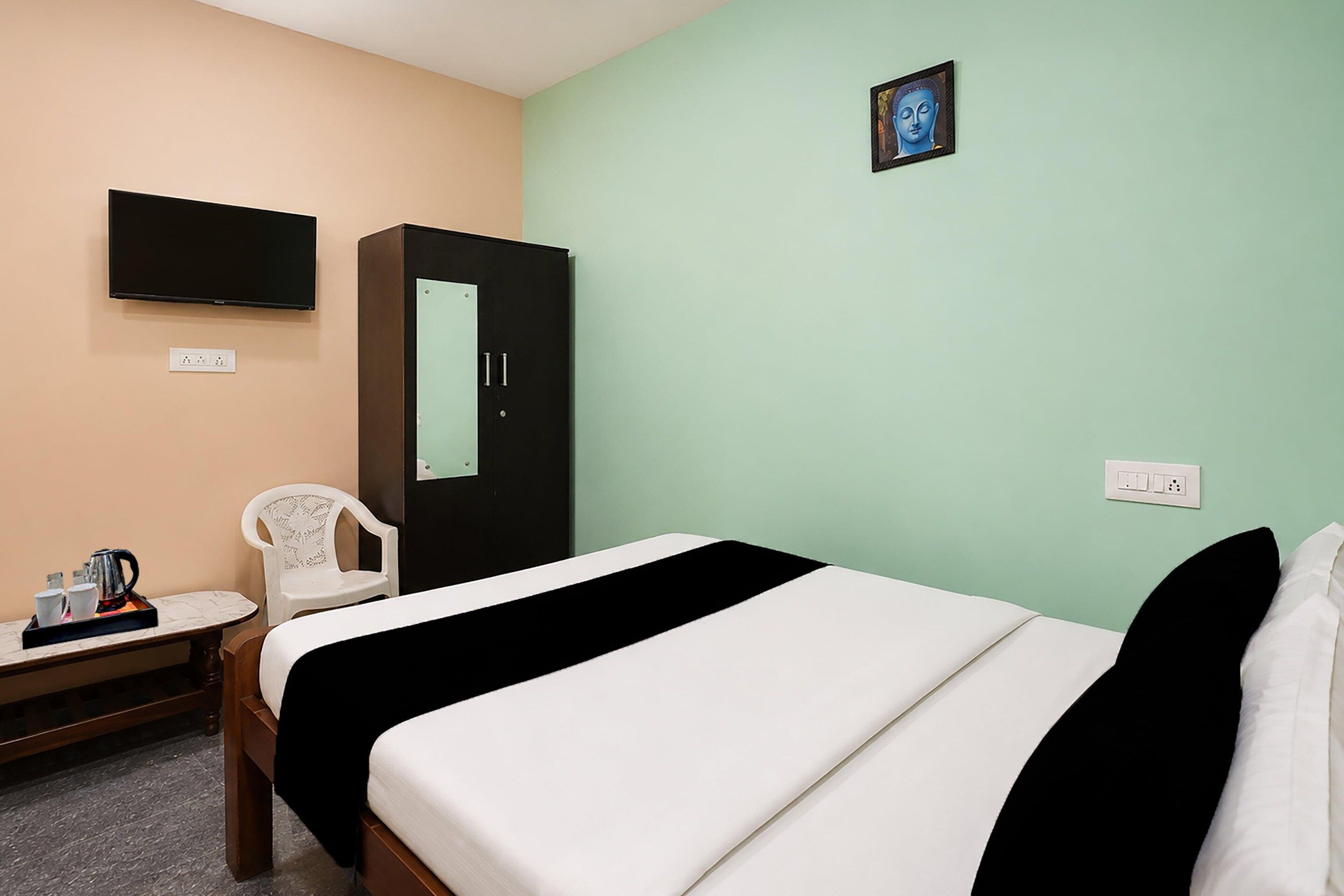 A hotel room with a bed, a desk with a kettle, a chair, a wardrobe, a TV, and a picture on the wall.