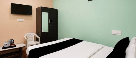 A hotel room with a bed, a desk with a kettle, a chair, a wardrobe, a TV, and a picture on the wall.