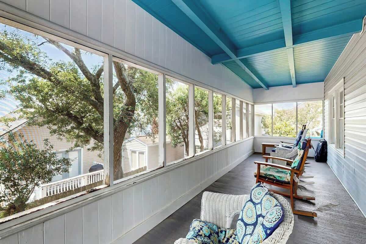 A covered porch with large windows, wicker furniture, and a view of trees and a house.
