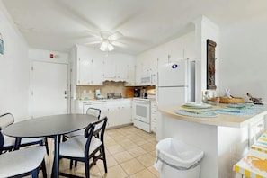 A compact kitchen with white cabinets, a microwave, a stove, and a dining area with a round table and chairs. - Beach Cottages at OD - 11B (North Myrtle Beach)