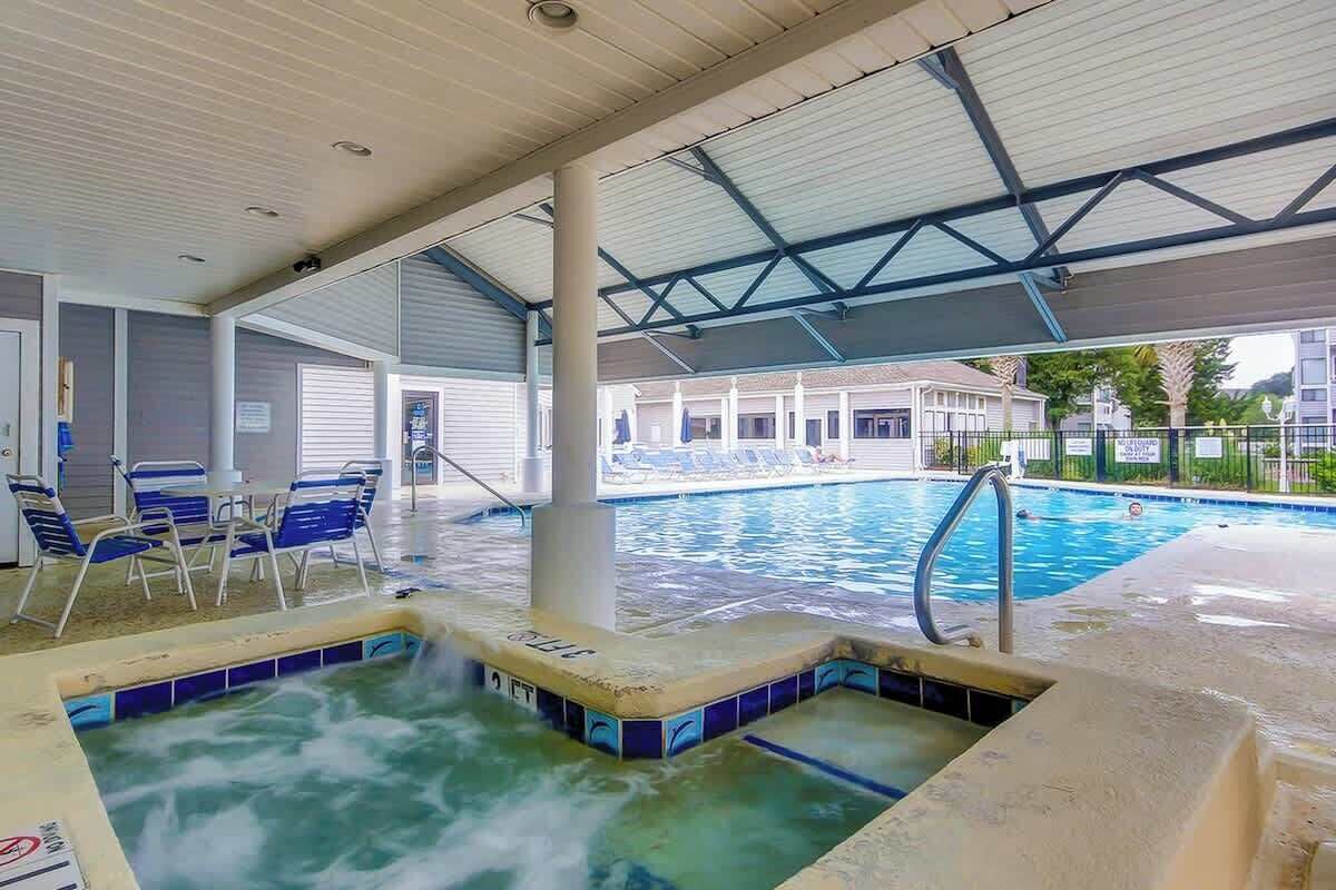 An indoor swimming pool with a hot tub, lounge chairs, and a covered area with a view of the pool.