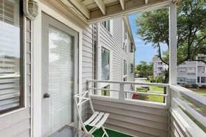 A covered porch with a white chair and a small table, a door with a window, and a view of neighboring houses. - Arcadian Dunes 12-245 (Myrtle Beach)