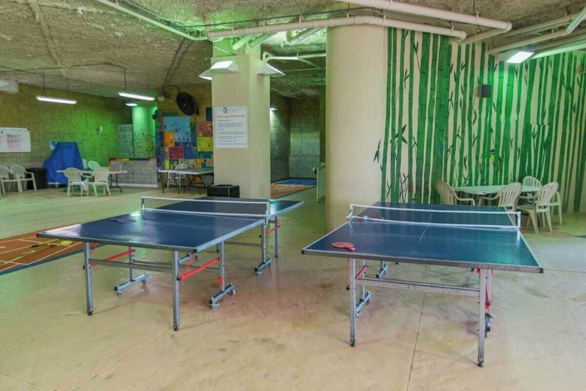 Ein Raum mit zwei Tischtennisplatten, einem Basketballfeld und einer grünen Wand mit vertikalen Linien.
