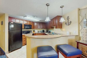 A kitchen with wooden cabinets, a black refrigerator, a microwave, a sink, and two blue bar stools. - Beachfront Bliss Retreat (Myrtle Beach)