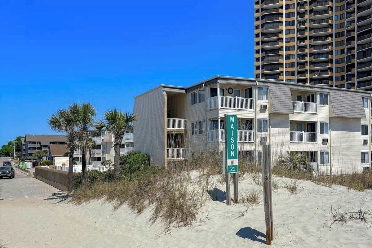 A beachfront building with balconies, a sign indicating the name 'MAISON 21', and a clear blue sky.