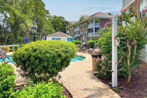 A pool area with a paved walkway, lounge chairs, and a building with balconies in the background. - Gardens at Cypress Bay 19-104 (Little River)