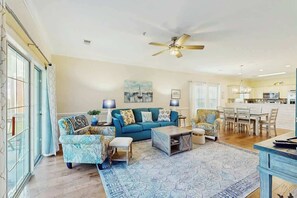 A spacious living room with a blue sofa, patterned armchairs, a coffee table, and a ceiling fan. - Gardens at Cypress Bay 19-104 (Little River)