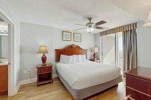 A hotel room with a large bed, wooden headboard, two bedside tables with lamps, a ceiling fan, and a large window with curtains. - Bahama Sands Unit 0903 (North Myrtle Beach)
