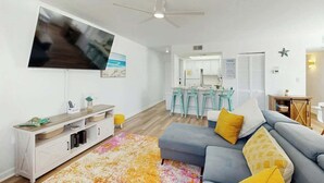 A modern living room with a flat-screen TV, a ceiling fan, a sofa with cushions, a coffee table, and a kitchen area in the background. - Dunes Pointe Unit B-10 (Myrtle Beach)