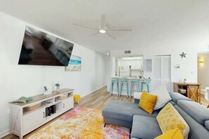 Ein modernes Wohnzimmer mit Flachbildfernseher, Deckenventilator, einer Couch mit Kissen, einem Couchtisch und einer Küchenzeile im Hintergrund.