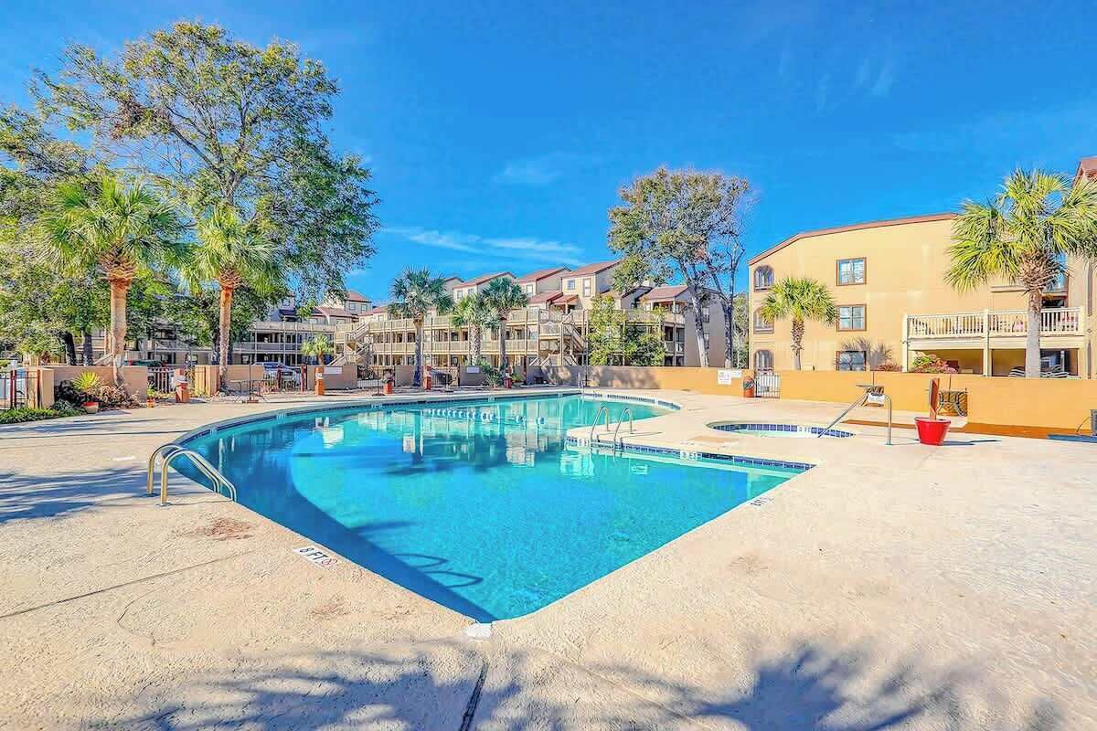 Piscina com superfície de água azul cristalina, rodeada por uma área arenosa com palmeiras e um edifício com varandas ao fundo.