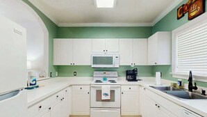 A kitchen with white cabinets, a microwave, an oven, a sink, and a window with blinds. - River Crossing Unit 124 (North Myrtle Beach)