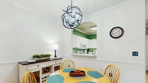 A dining area with a round table, four chairs, a chandelier, a clock, and a mirror reflecting a kitchen area. - River Crossing Unit 124 (North Myrtle Beach)
