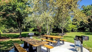 An outdoor picnic area with concrete tables and benches, surrounded by trees and grass. - River Crossing Unit 124 (North Myrtle Beach)