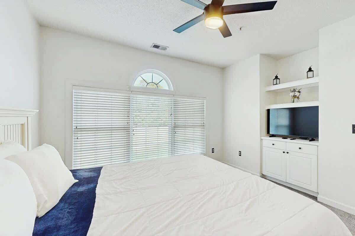 Ein Schlafzimmer mit einem Bett, einem Deckenventilator, einem Fenster mit Jalousien und einem Fernseher auf einem weißen Schrank.