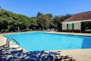 A clear blue swimming pool with a 5 FT depth limit sign, surrounded by trees and a building with a tiled roof. - River Crossing Unit 124 (North Myrtle Beach)