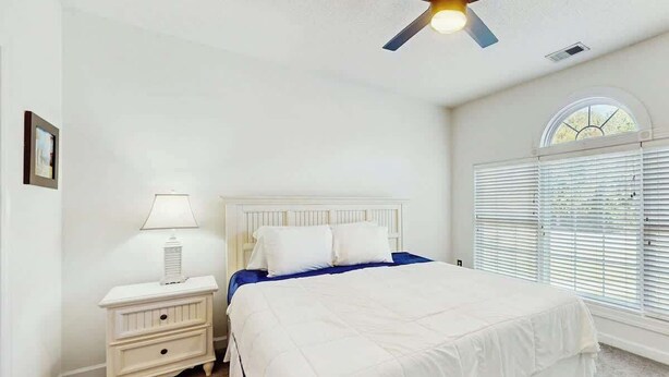 A bedroom with a bed, nightstand, lamp, ceiling fan, and a window with blinds. - River Crossing Unit 124 (North Myrtle Beach)
