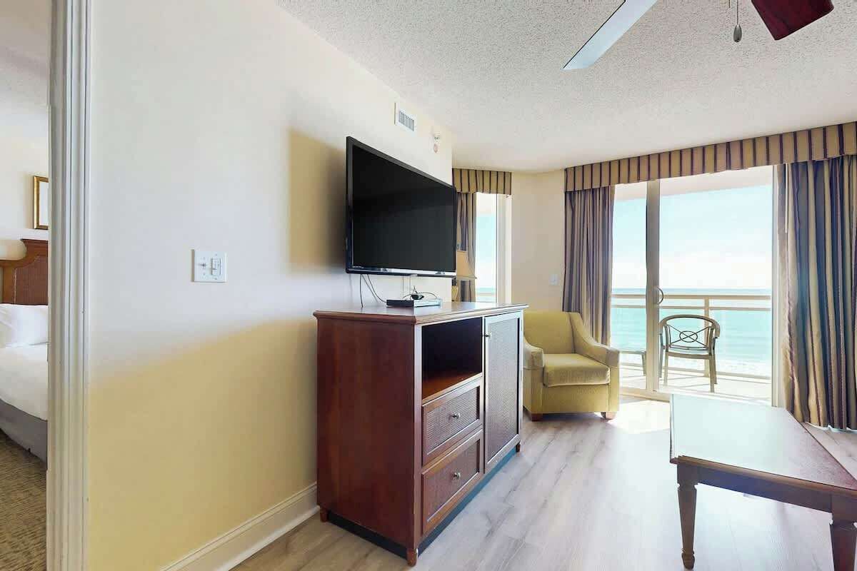 A hotel room with a television on a wooden cabinet, a yellow armchair, a dining table, and a view of the ocean through large windows.