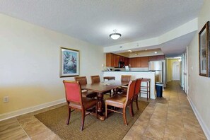 A dining area with a wooden table and chairs, a kitchen in the background, and a framed picture on the wall. - Bahama Sands Unit 0603 (North Myrtle Beach)