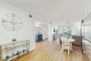 A modern apartment with a kitchen, dining area, and living room. There is a large wall clock, a glass coffee table, and a sofa. - Sea Cabin Unit 137 Condo (North Myrtle Beach)
