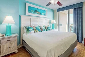 A bedroom with a bed, two bedside tables, a ceiling fan, a painting, and a view of the outdoors. - Bahama Sands Unit 1107 (North Myrtle Beach)