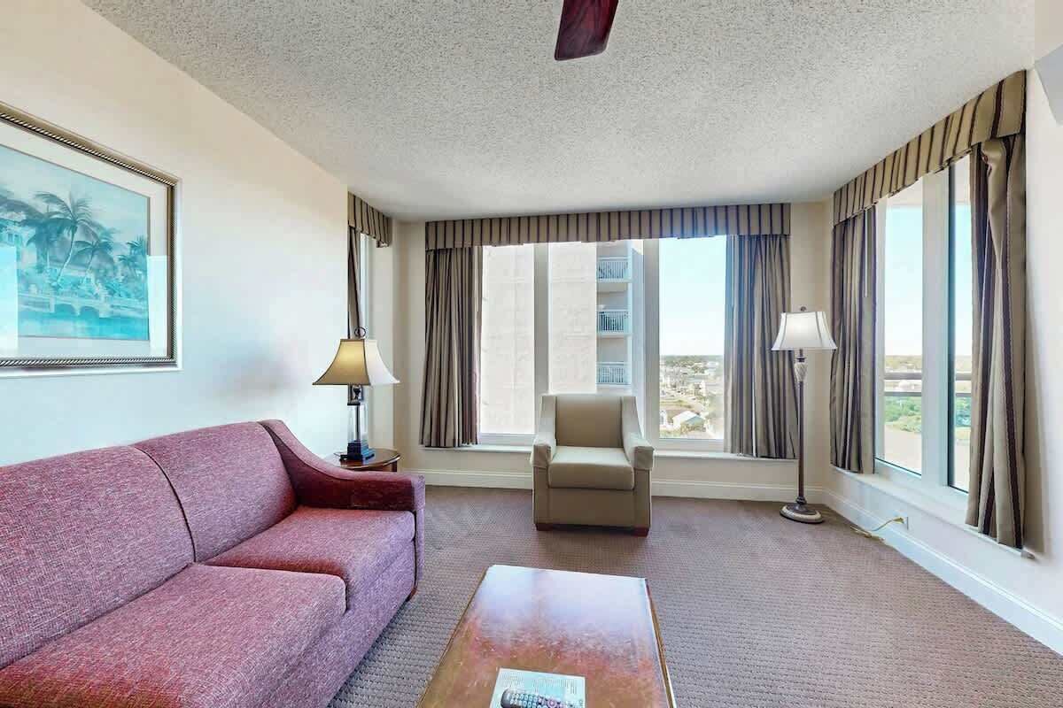 A hotel room with a red sofa, a wooden coffee table, a chair, and a large window with curtains.