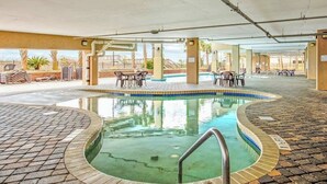 Ein Hallenbad mit gefliestem Deck, umgeben von Liegestühlen und Tischen, mit Blick auf Palmen und ein Gebäude im Hintergrund.