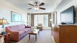 Ein Hotelzimmer mit Deckenventilator, Fernseher, einer Couch, einem Couchtisch, einem Sessel und einem großen Fenster mit Vorhängen.