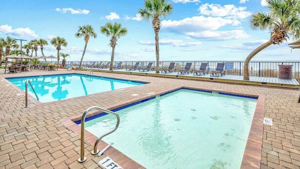 A pool area with two swimming pools, palm trees, lounge chairs, and a clear sky. - Compass Cove Unit 560 (Myrtle Beach)