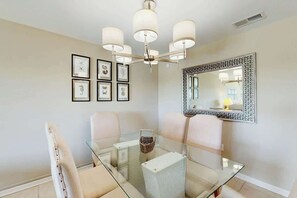 A dining area with a glass table, white chairs, a chandelier, framed artwork, and a decorative mirror. - Tides Unit C-1 Condo (North Myrtle Beach)