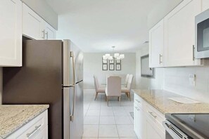 A modern kitchen with white cabinetry, a stainless steel refrigerator, a dining area with a table and chairs, and a built-in oven. - Tides Unit C-1 Condo (North Myrtle Beach)