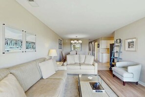 A modern living room with a sofa, a coffee table, and a TV. There is a dining area with a table and chairs in the background. - Tides Unit C-1 Condo (North Myrtle Beach)