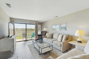 A modern living room with a sofa, coffee table, and a TV. There is a sliding glass door leading to an outdoor area. - Tides Unit C-1 Condo (North Myrtle Beach)