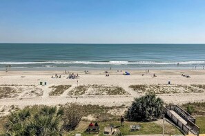 Ein Strand mit einer Holzpromenade, die zu einer kleinen Struktur führt, und Menschen, die am Sand entspannen.