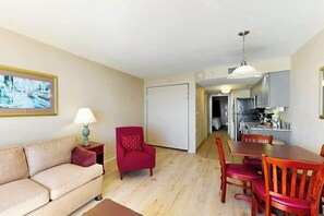 Ein Wohnzimmer mit einer Couch, einem roten Sessel, einem Esstisch mit Stühlen und einer Küche im Hintergrund.