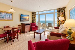 A hotel room with a dining area, a sofa, a red armchair, a table, and a view of the ocean. - Oceanfront Luxe | Atlantica Seven Fifty-Five (Myrtle Beach)