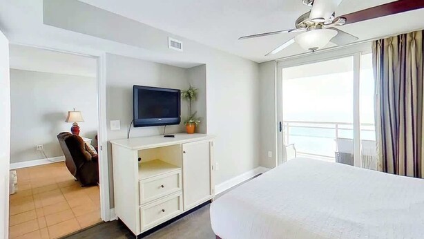 A hotel room with a bed, a television on a cabinet, a ceiling fan, and a view of the sea through large windows. - Bahama Sands Unit 0502 (North Myrtle Beach)