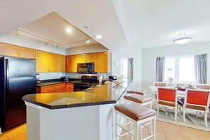 A modern kitchen with a central island, a dining area with red chairs, and a large window providing natural light. - Bahama Sands Unit 0502 (North Myrtle Beach)