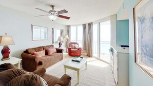 A living room with a ceiling fan, a brown sofa set, a red armchair, a white coffee table, and a large window with curtains. - Bahama Sands Unit 0502 (North Myrtle Beach)