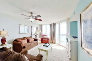 A living room with a ceiling fan, a brown sofa set, a red armchair, a white coffee table, and a large window with curtains. - Bahama Sands Unit 0502 (North Myrtle Beach)