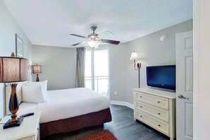 A hotel room with a bed, a television, a desk with a lamp, a ceiling fan, and a window with curtains. - Bahama Sands Unit 0502 (North Myrtle Beach)
