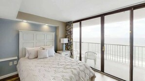 A bedroom with a large bed, a nightstand, a lamp, a window with a view, and a balcony with a chair. - Sands Ocean Club 1629/1630 (Myrtle Beach)