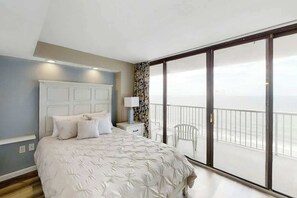 A bedroom with a large bed, a nightstand, a lamp, a window with a view, and a balcony with a chair. - Sands Ocean Club 1629/1630 (Myrtle Beach)