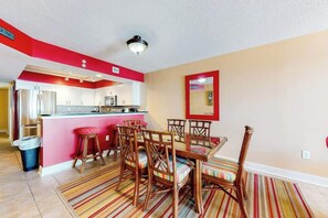 A compact kitchen with a central island, a dining area with wooden chairs, and a striped rug on the floor. - Bahama Sands Unit 0506 (North Myrtle Beach)