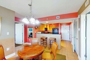 A dining area with a wooden table and chairs, a kitchen with a central island, and a hallway leading to a door. - Bahama Sands Unit 1201 (North Myrtle Beach)