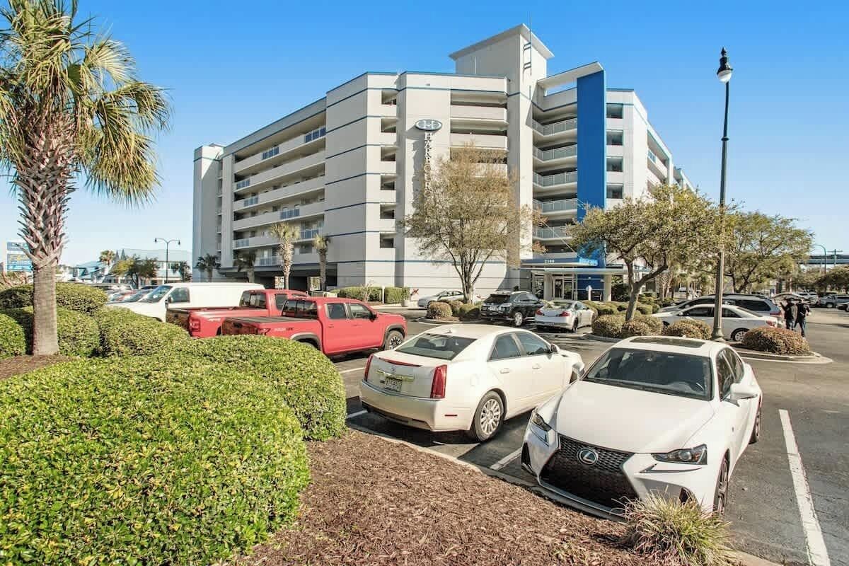 A multi-story parking garage with several cars parked, surrounded by greenery and a clear sky.